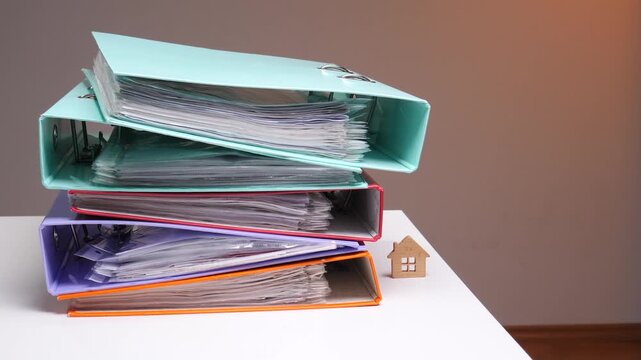 Person holding colorful ring binders of important property documents next to a small wooden house model on a white desk, symbolizing real estate ownership, mortgage and finance paperwork