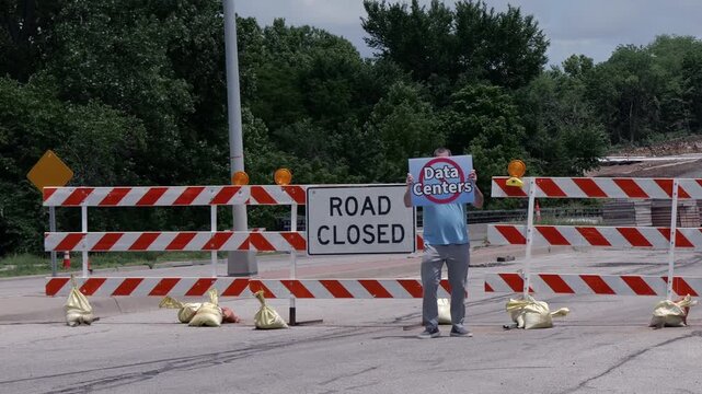 Man holding No Data Centers sign construction site barricades protesting AI tech infrastructure power grid development environmental impact zoning