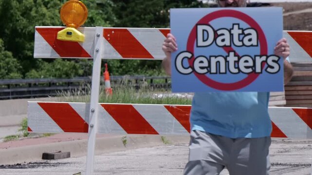 Man holding No Data Centers sign construction site barricades protesting AI tech infrastructure power grid development environmental impact zoning
