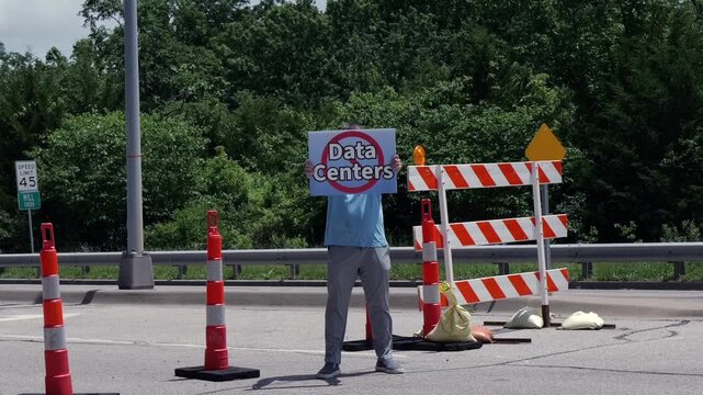 Man holding No Data Centers sign construction site barricades protesting AI tech infrastructure power grid development environmental impact zoning