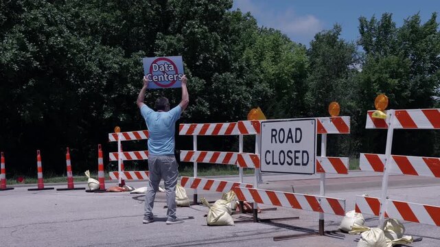 Man holding No Data Centers sign construction site barricades protesting AI tech infrastructure power grid development environmental impact zoning