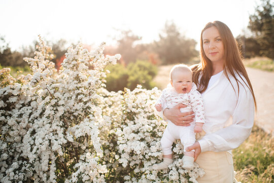 Young woman 30-35 year old holding crying baby over flowers in sunny day outdoors. Motherhood.