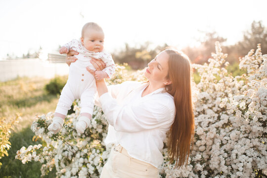 Cheerful young woman 30-35 year old holding baby over flowers in sunny day outdoors. Summer vacation season. Motherhood.