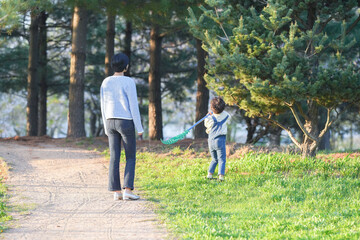Obraz premium Sunlight bathes a quiet park path. Woman and child walk side by side. The child holds a kite, eyes wide with joy. Trees loom softly in the background. Grass glows green beneath their feet