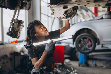 African american mechanic working in an auto repair garage, looking at a lifted car's undercarriage and using a light © anatoliycherkas
