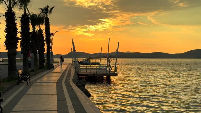 A golden sunset over the sea in Gulluk, Turkey. A coastal promenade with palm trees, a small wooden pier, and distant dark hills.