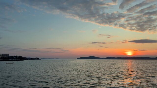 A calm, warm sunset over the Aegean Sea in Gulluk, Turkey. The sun hangs low in a hazy orange sky, casting a bright golden light path across the gently rippling water toward distant dark hills.