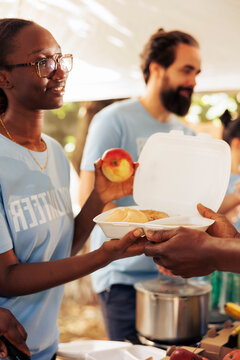 Close-up shot of generous black woman working in community food bank, serving free meals for the needy. Volunteering team of charity workers helping and sharing nutritrious items to homeless people.