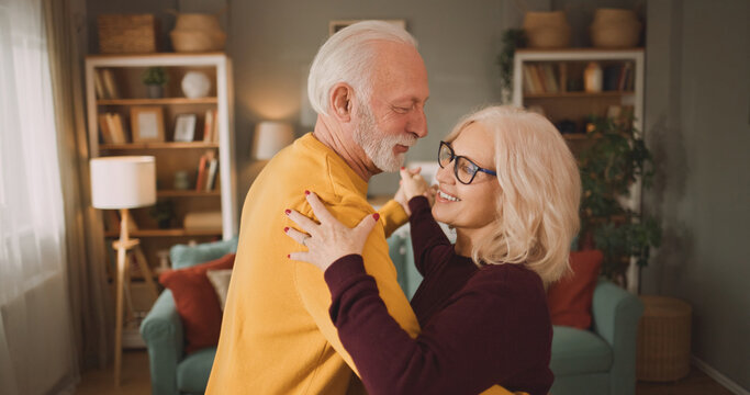 An elderly couple enjoys dancing together in their living room. They hold each other close and smile while soft sunlight comes through the window.