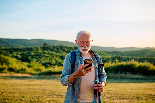 Senior man smiling, using a mobile phone with trekking poles in hand, standing in nature with hills in the background