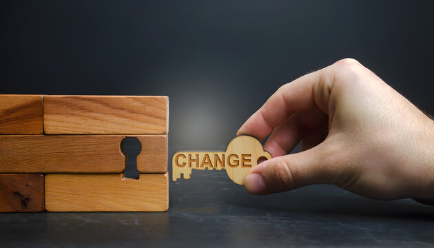 A hand holding a wooden key with the word Change written on it, moving towards a keyhole shaped gap in a stack of wooden blocks against a dark background, conceptual image.