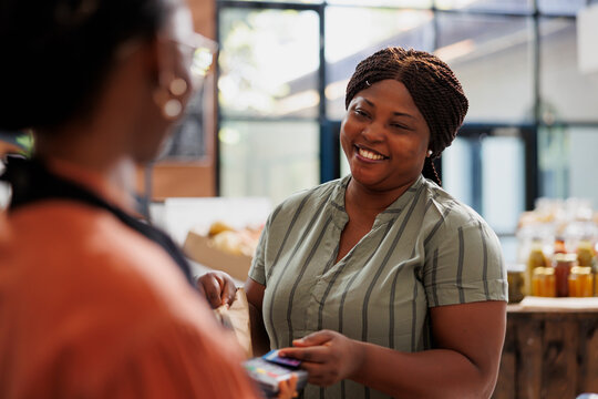 Female customer buys fresh, healthy groceries and pays with debit card. At counter with different products, African American merchant holds POS for black woman to make payment.
