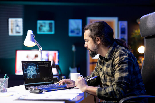 Male programmer coding for AI and machine learning on digital laptop, bearded man looking at neural network and database on his electronic device while comparing with information on his notebook.