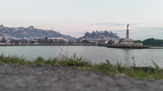 Cinematic low-angle ground shot of Lake Anosy and the Monument aux Morts with the Antananarivo skyline in the background during dusk in Madagascar.