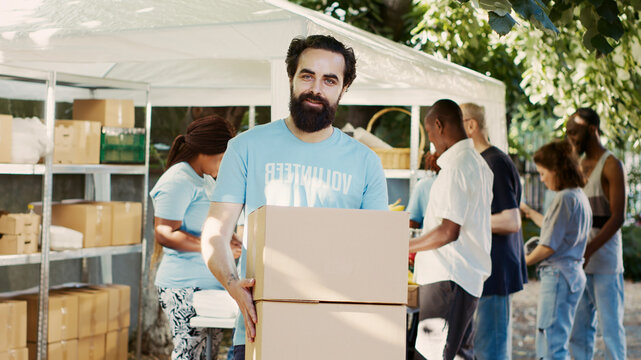 Generous man volunteering at neighborhood food drive, distributing non-perishable rations to the hungry and homeless, while holding donation box, caucasian charity worker looks at camera.