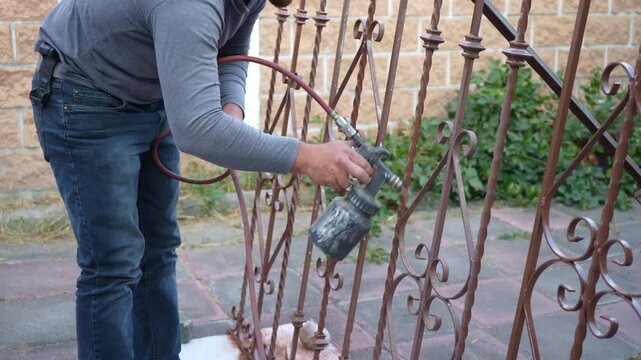 Worker spray painting metal window bars with an air compressor spray gun. Metalwork maintenance, corrosion protection, steel preservation, protective coating process