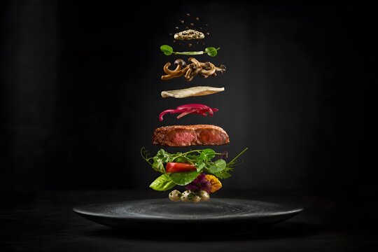 Gourmet steak with floating ingredients stacked above a black plate. Creative culinary composition of meat, mushroom, vegetable and herb. Fine dining food concept for restaurant menu.