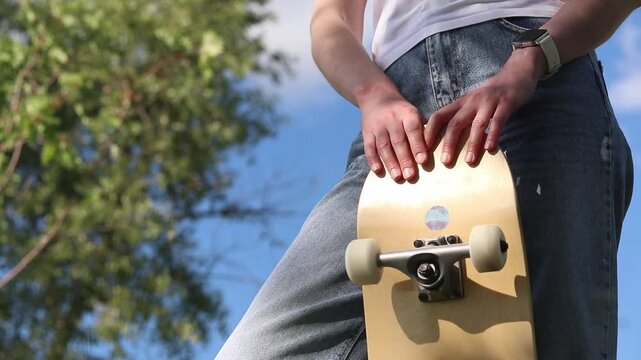 Young girl in white t-shirt and jeans holding skateboard against blue sky and trees. Bottom view. Stylish skateboarder girl posing with her board outdoors on a sunny day. Active leisure concept