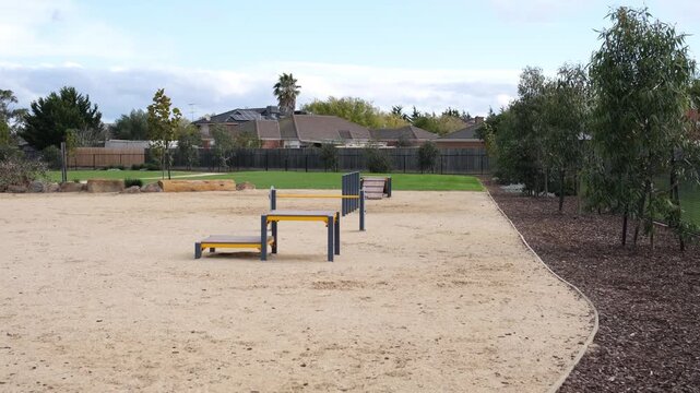 An off-leash dog park featuring sandy ground surface, dog agility and obstacle equipment bordered by a mulched native garden bed, in a suburban residential neighbourhood in Melbourne, Australia.