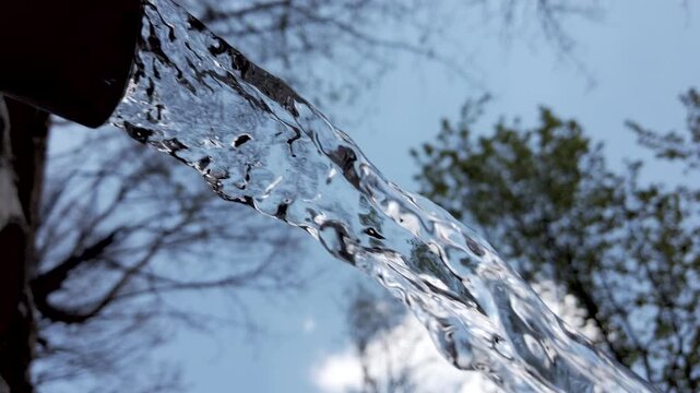 Water flows from a spout into a stream under a cloudy sky in the afternoon near trees in a natural setting during springtime