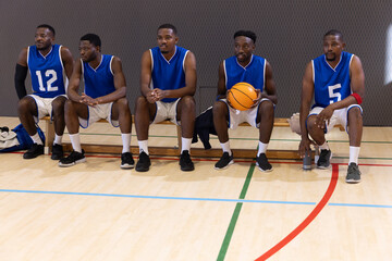 Naklejka premium African American male teammates sitting on bench in gym in blue jerseys holding orange basketball
