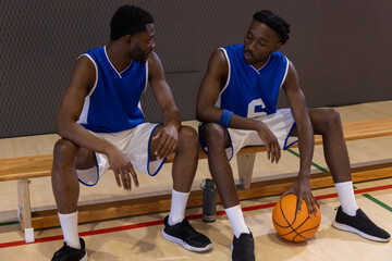 Fototapeta premium Two African American men sitting on gym bench wearing blue jerseys holding orange basketball