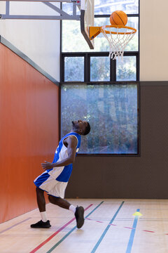 Orange basketball is passing through rim above glass backboard on wooden court near window