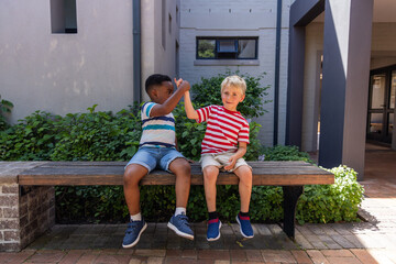 Naklejka premium Diverse boys high-fiving while sitting on wooden bench in sunny brick courtyard with sneakers