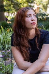 Fototapeta premium African American woman sitting on low bench in garden, wearing black top, gazing upward right