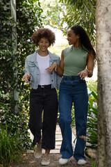 Obraz premium African American female friends walking and talking along garden path with trellis and denim jacket