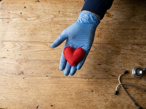 Doctors gloved hand holding a small red heart on a wooden table with a stethoscope.