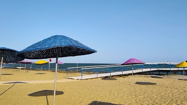 Empty Sandy Beach Resort with Colorful Woven Straw Sun Umbrellas and a Wooden Walking Pier Leading into the Sea Under Clear Blue Sky