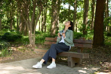 Fototapeta premium Young woman with camera relaxing on bench in green park