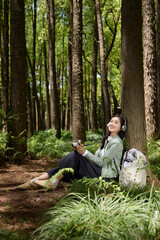 Fototapeta premium Young woman with camera and headphones resting under tree in forest