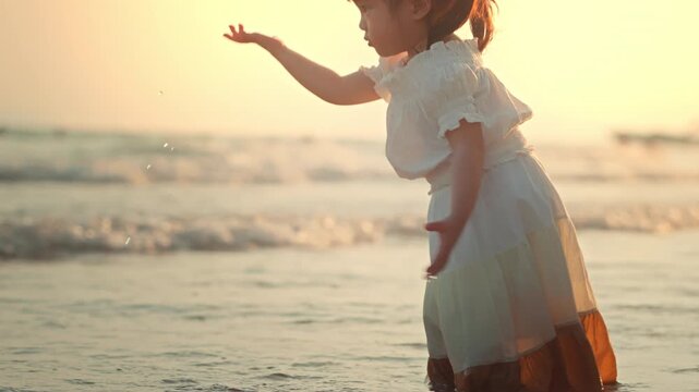 Cute little Asian girl in dress playing with sea water on tropical beach at sunset, happy curious child enjoying summer vacation and exploring nature during golden hour for family travel and parenting