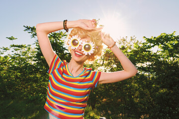 Obraz premium Cheerful woman outdoors, wearing a striped shirt and sunflower sunglasses, basking in warm sunshine, carefree pose, vibrant colors, lush greenery background, joyful spontaneous moment, lively