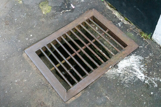 Rusted metal rainwater drain cover installed in a concrete sidewalk. Its rectangular design features parallel iron bars to allow water to drain away.