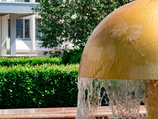 Water cascades smoothly over a large, golden dome-shaped fountain set in a courtyard surrounded by green hedges and residential buildings. Sunlight highlights the reflective surface of the water and t © Yarr