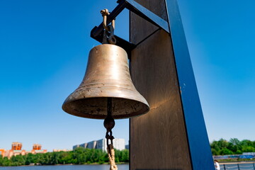 Fototapeta premium Bronze bell suspended from a dark metal frame with a rope clapper inside, positioned outdoors near a body of water. Clear blue sky contrasts with greenery and urban buildings across the water in the b