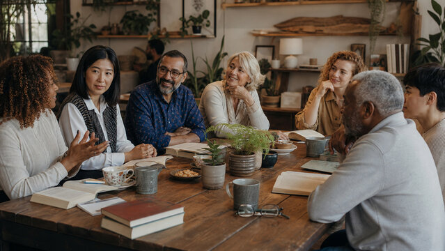 Diverse group of adults sharing a thoughtful book discussion around a wooden table in a warm cafe with plants, mugs, and open books.