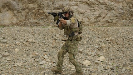 Obraz premium A special forces soldier in full tactical gear patrols through a rocky quarry, aiming his assault rifle during a military training operation or combat mission.