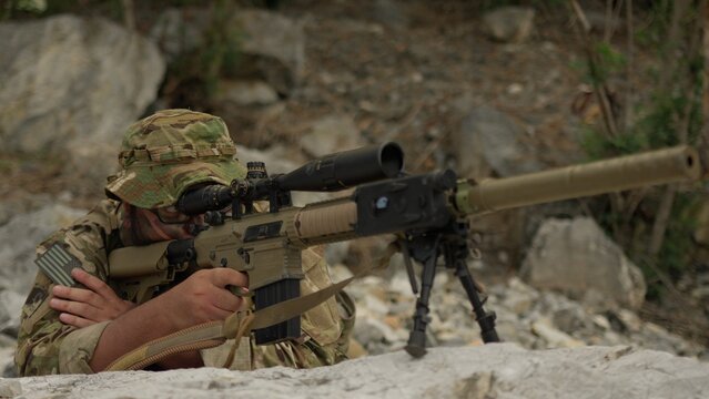 A military sniper in camouflage lies in a concealed position, aiming his advanced rifle with a scope during a tactical surveillance operation in the mountains.