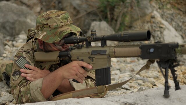 A military sniper in multicam camouflage aims his rifle from a concealed rocky position, conducting surveillance during a special operations mission.