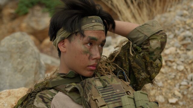 A young Asian soldier with face paint in full tactical gear remains vigilant while on a reconnaissance mission in a rugged, hostile outdoor environment.
