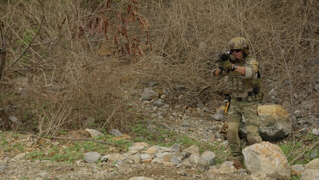 A special forces operator in full combat gear secures a perimeter while patrolling through rugged, hostile wilderness terrain for a military operation.