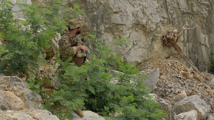 Obraz premium A soldier in full tactical gear patrols a rugged mountain area, conducting surveillance and maintaining security during a special operations reconnaissance mission.
