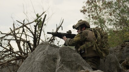 Obraz premium A special forces soldier in full tactical gear aims an assault rifle from behind a rock, conducting surveillance during a covert military operation outdoors.