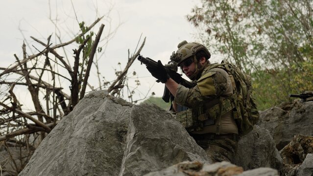 A special forces operator in full combat gear takes a defensive position, aiming an assault rifle from behind large rocks during a tactical reconnaissance mission.