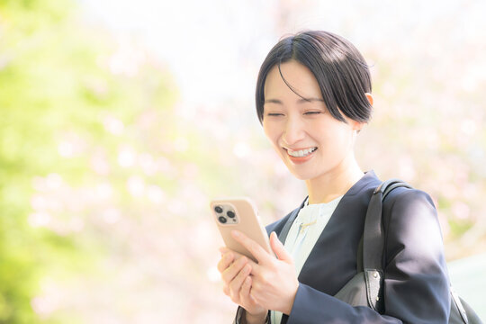 スマートフォンを見る笑顔の女性のビジネスシーン春の桜の明るい通勤や風景イメージ