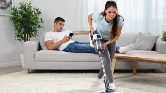 Frustrated woman vacuuming carpet with cordless vacuum cleaner, lazy husband relaxing on sofa with smartphone, domestic conflict and unequal housework